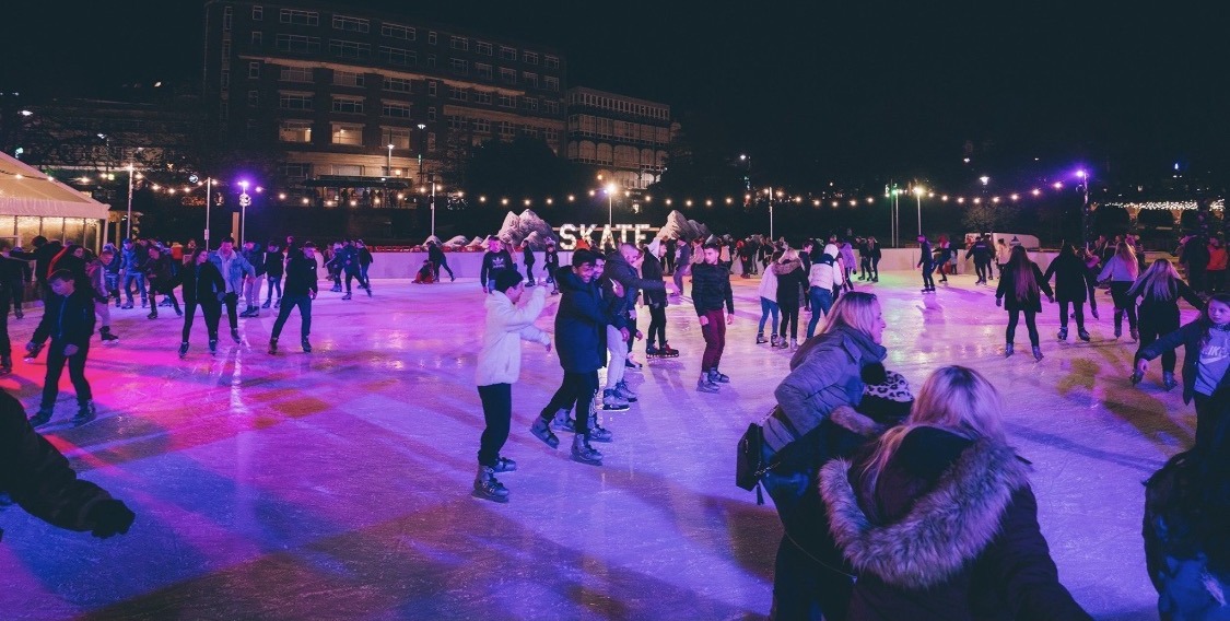 Skaters gliding on the outdoor ice rink in Bournemouth at night, beneath rows of festoon lights and a lit Skate sign